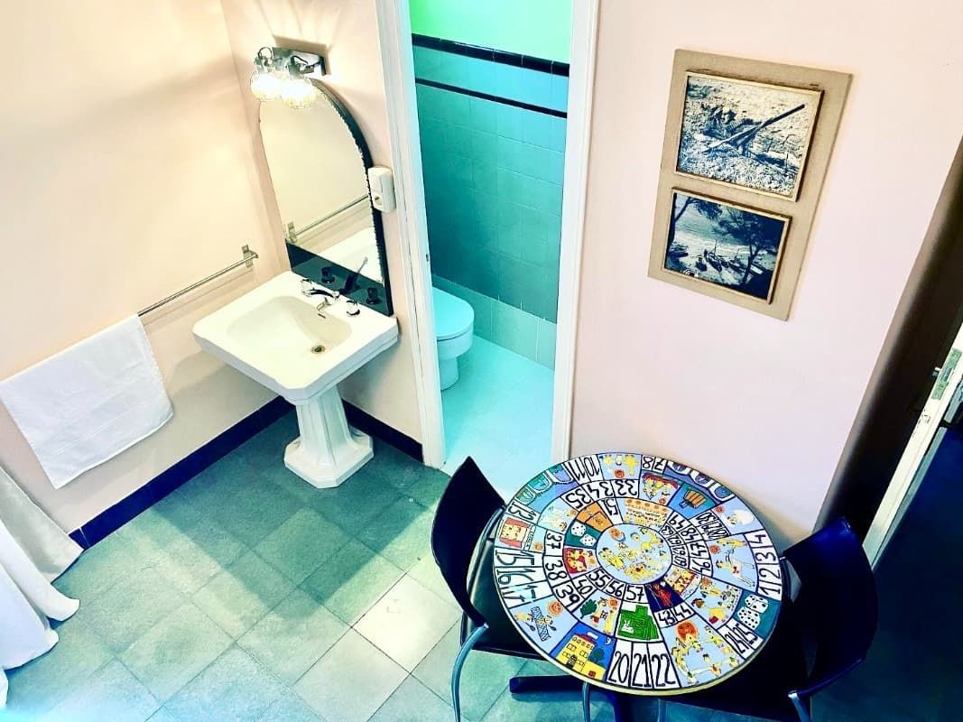 Baño pequeño con lavabo de pedestal, ducha y mesa redonda de mosaico con sillas negras.