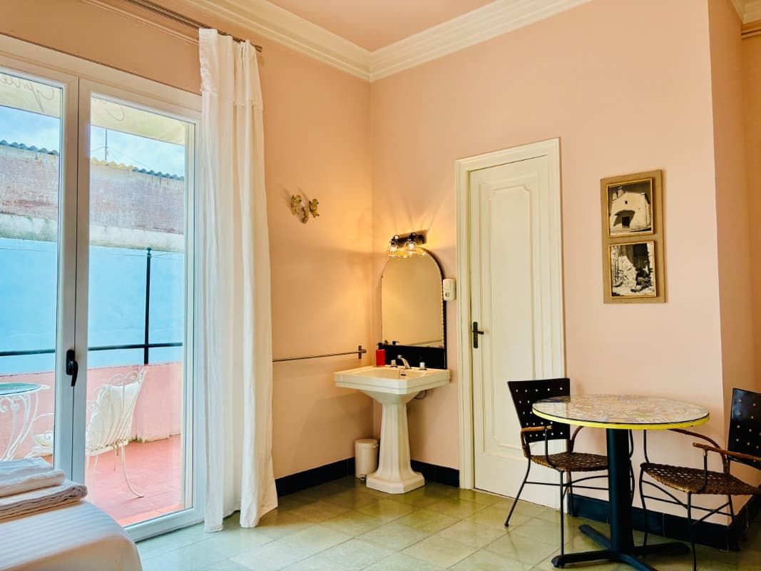 Habitación rosa con puerta de cristal, lavabo blanco y mesita junto a un armario con espejo.