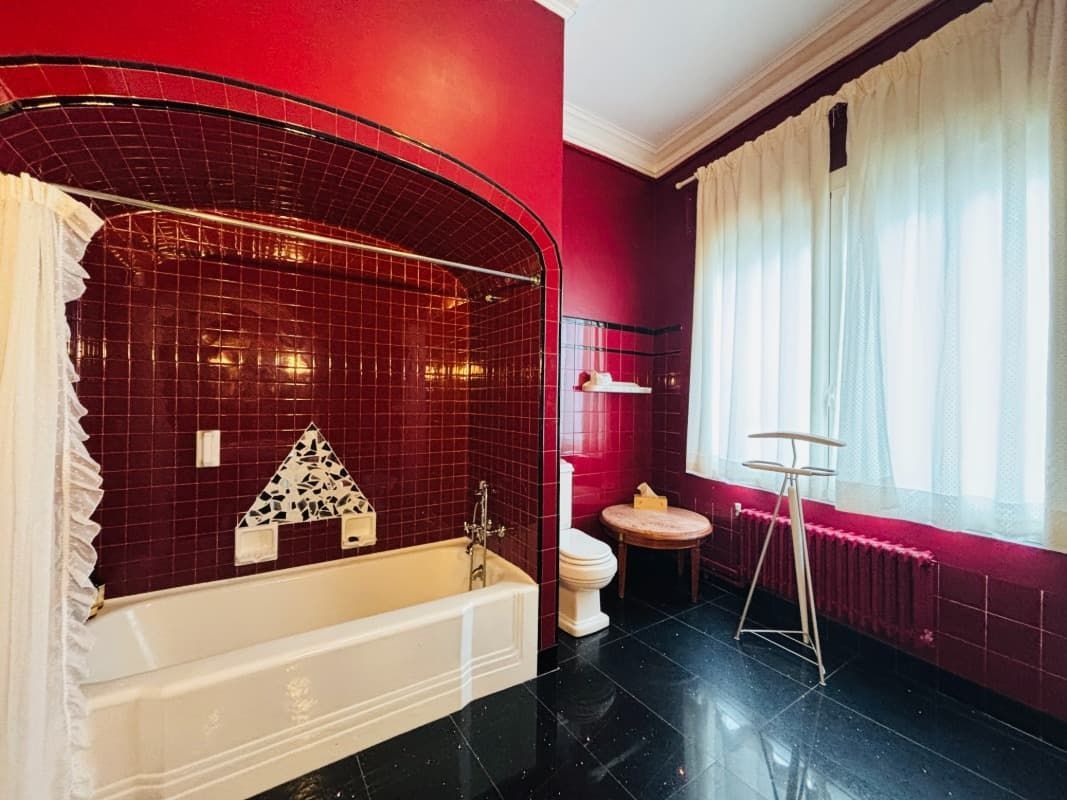 Elegante baño con paredes color burdeos, bañera blanca, suelo de baldosas oscuras y una ventana luminosa.