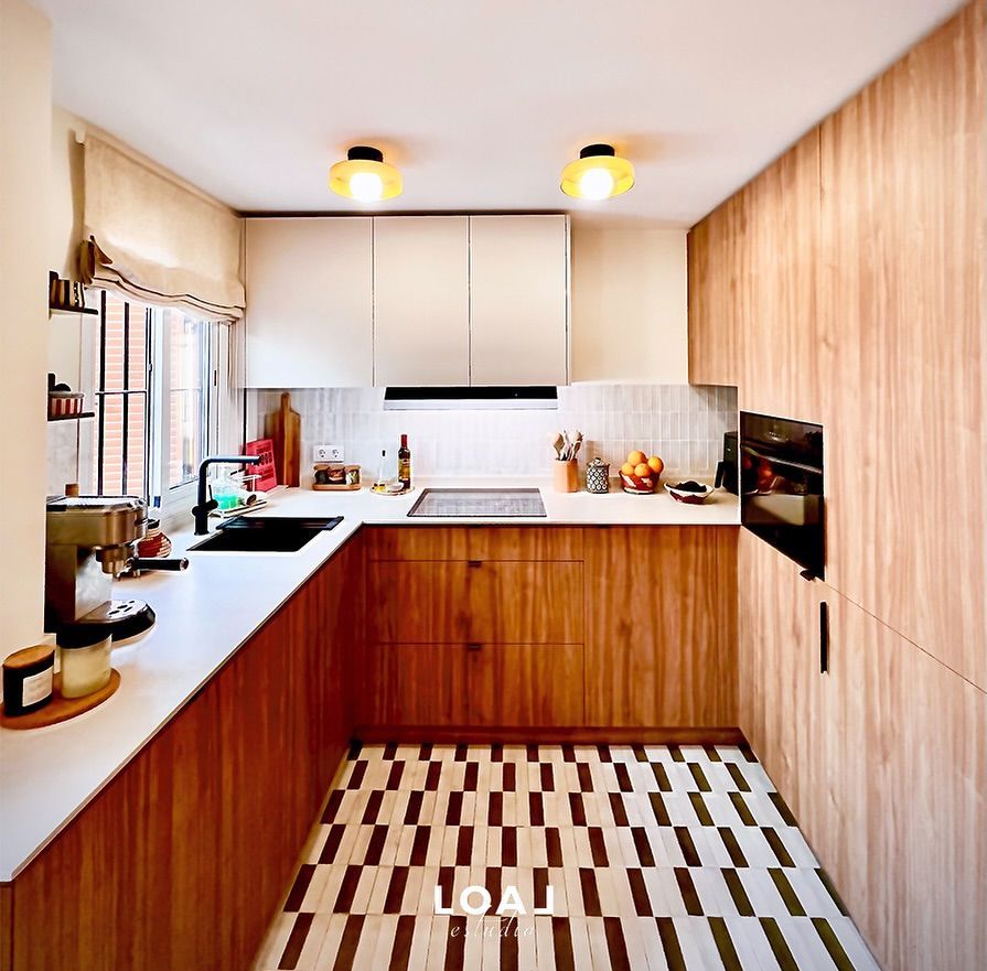 Cocina moderna y luminosa con armarios de madera, encimeras blancas, suelo estampado y dos lámparas de techo