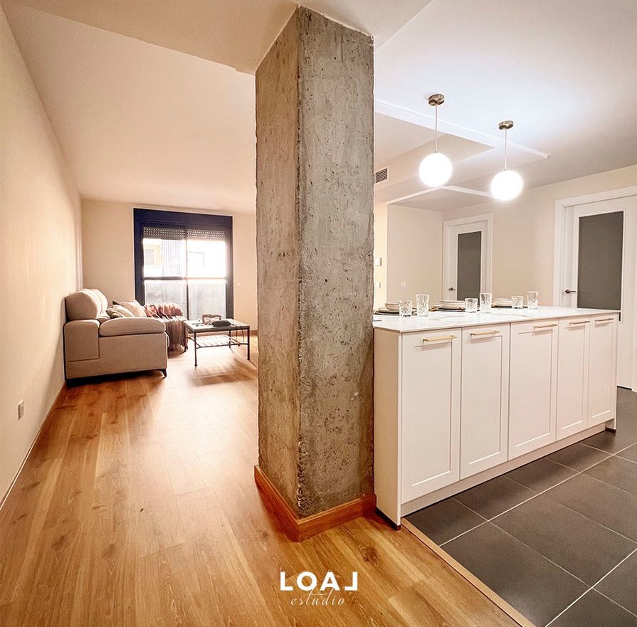 Apartamento diáfano con isla de cocina, columna de hormigón, suelos de madera y un sofá junto a una ventana luminosa