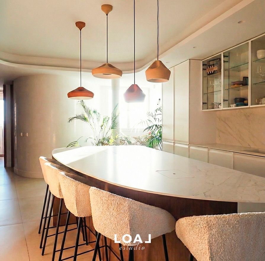 Comedor moderno con una mesa larga, sillas de mimbre, lámparas colgantes y una ventana luminosa con plantas