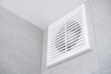 Grille d'aération blanche pour salle de bains, près du plafond, sur un mur carrelé gris clair.