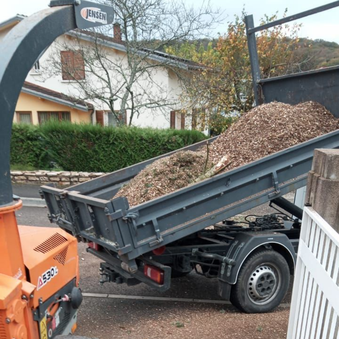 Petite remorque chargée de paillis à côté d'un broyeur de branches d'oranger dans une cour résidentielle.