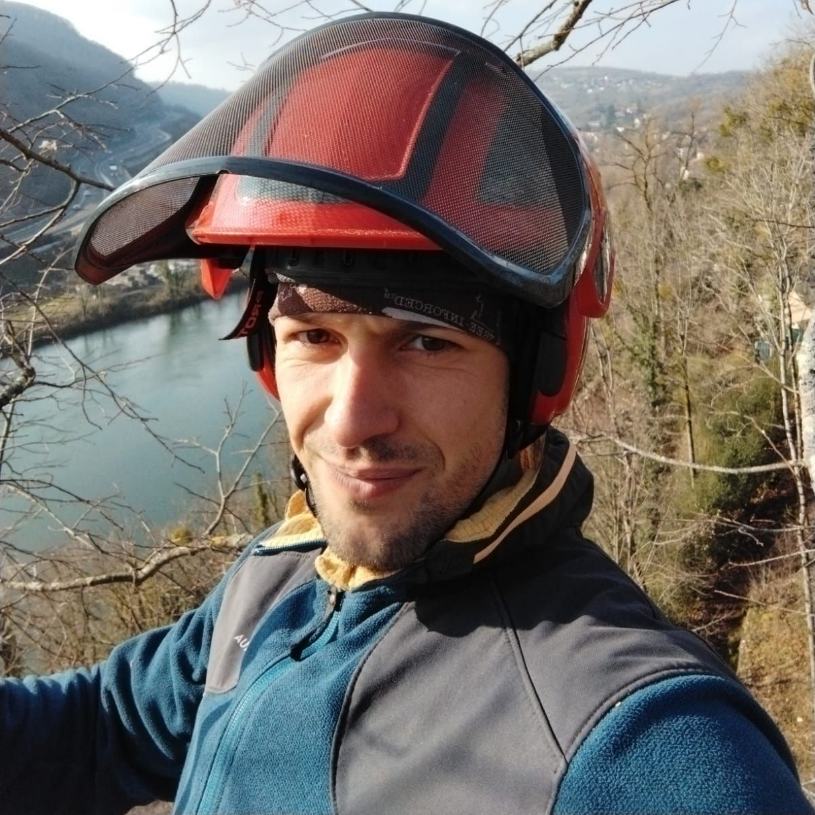 Personne souriante portant un casque rouge et une veste bleue sur une colline surplombant une rivière