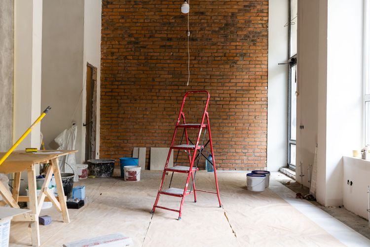 Habitación parcialmente reformada con pared de ladrillo visto, escalera roja y herramientas de construcción.