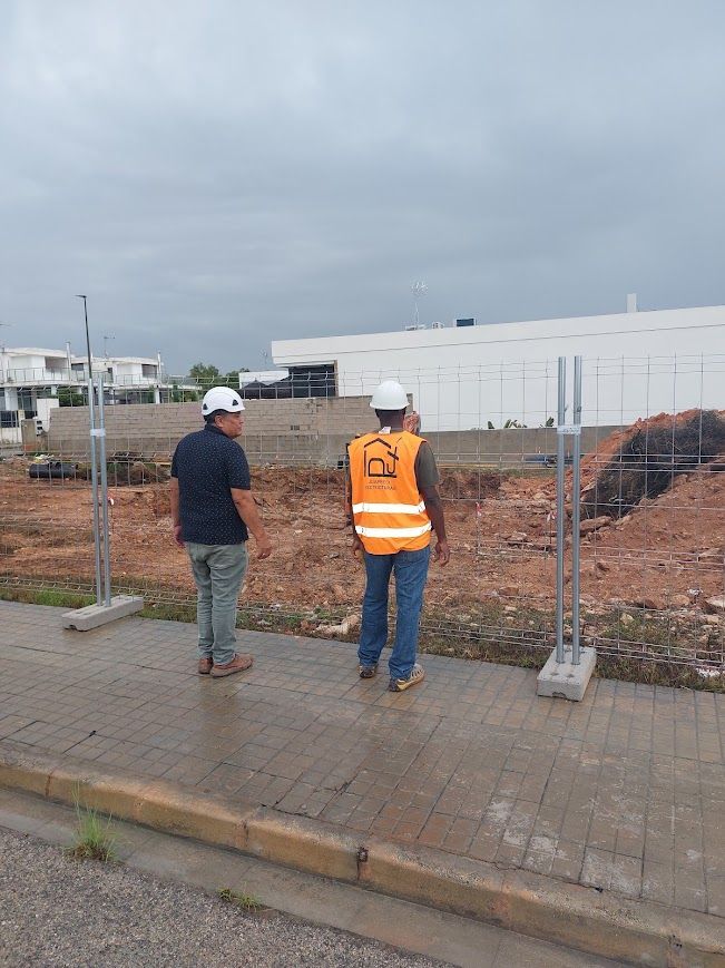 Dos obreros de la construcción con cascos de seguridad observan una excavación vallada bajo un cielo nublado.
