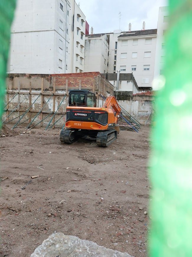 Excavadora naranja en una obra en construcción embarrada junto a altos edificios blancos.