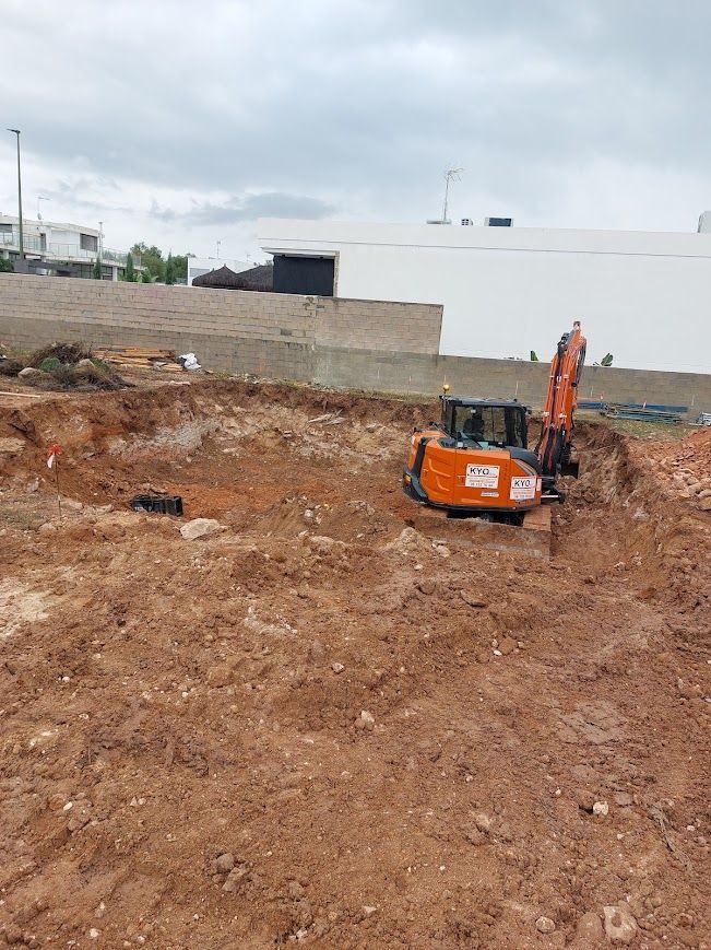 Excavadora naranja en una obra de construcción de tierra junto a un edificio blanco y un muro de bloques 