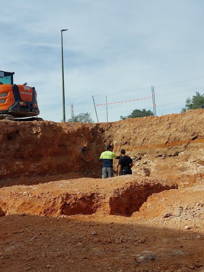 Dos trabajadores se encuentran en una gran excavación de tierra junto a una excavadora naranja en una obra de construcción.