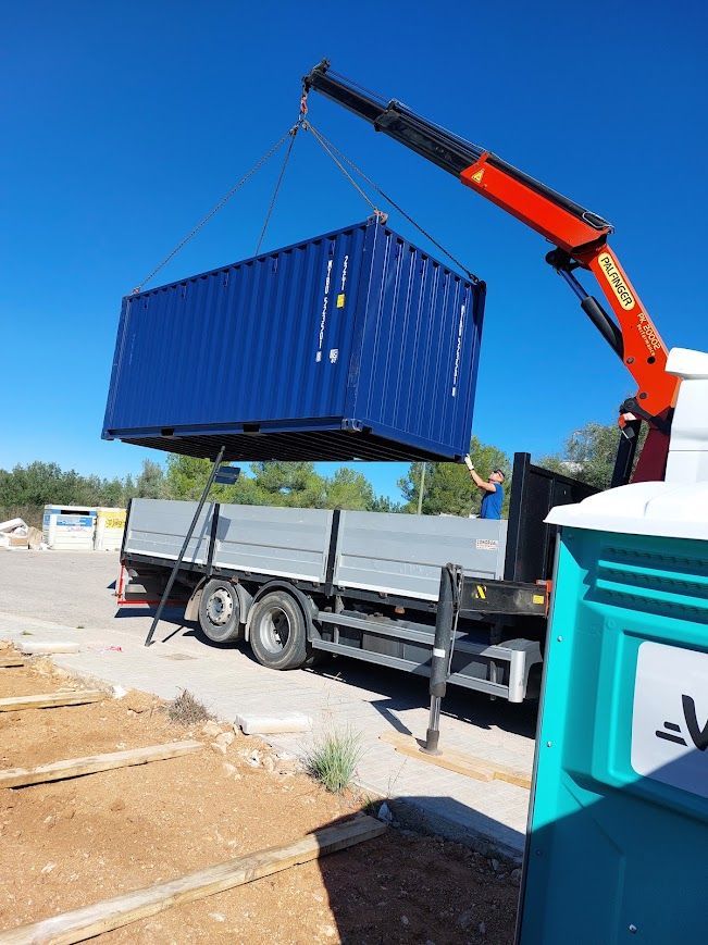 Una grúa naranja levanta un contenedor de transporte azul sobre un camión de plataforma en un patio industrial.
