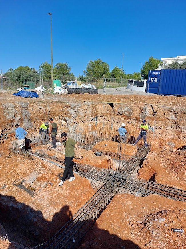 Equipo de construcción trabajando en una gran excavación con barras de refuerzo y zanjas bajo un cielo azul despejado.