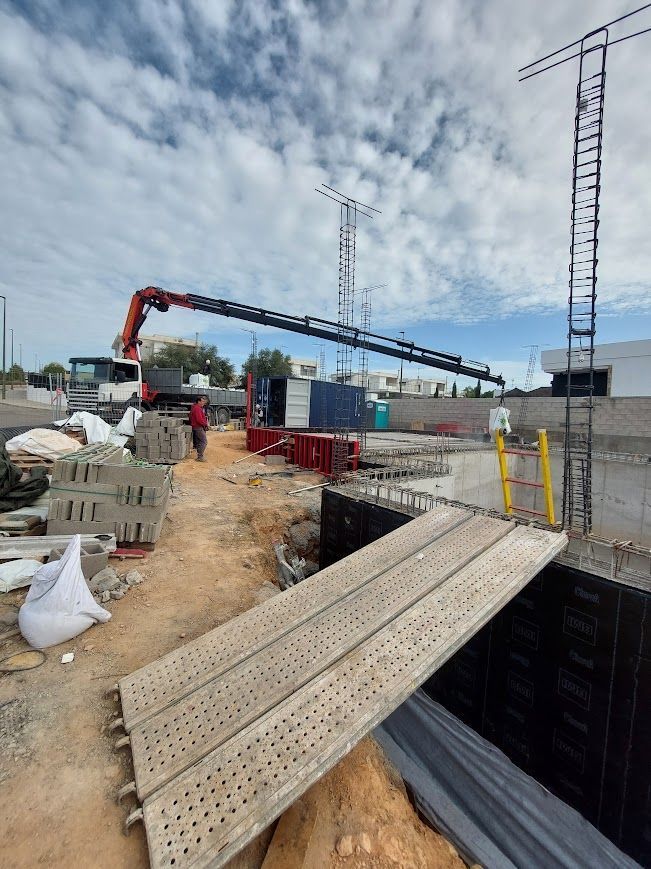 Obra en construcción con grúa, trabajadores y un puente de tablones de madera sobre una excavación.