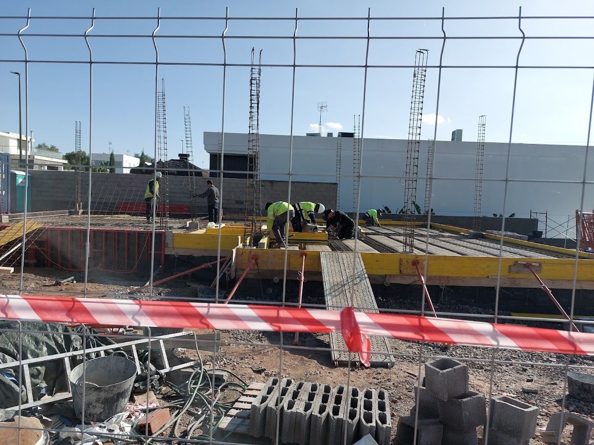 Obra en construcción con trabajadores en un tejado parcialmente construido, vista a través de una valla de seguridad.