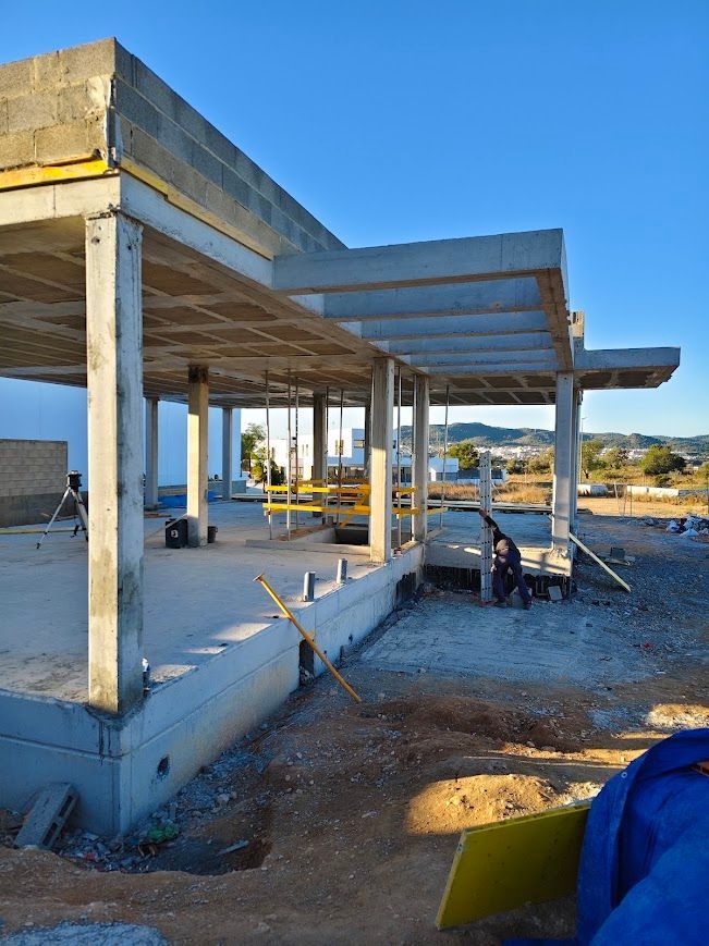 Casa de hormigón parcialmente construida con columnas y vigas de techo a la vista en un solar soleado.