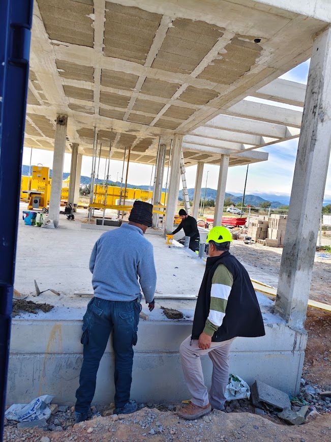 Obreros en una obra de construcción de hormigón bajo un techo parcialmente terminado, con una persona de pie en primer plano.