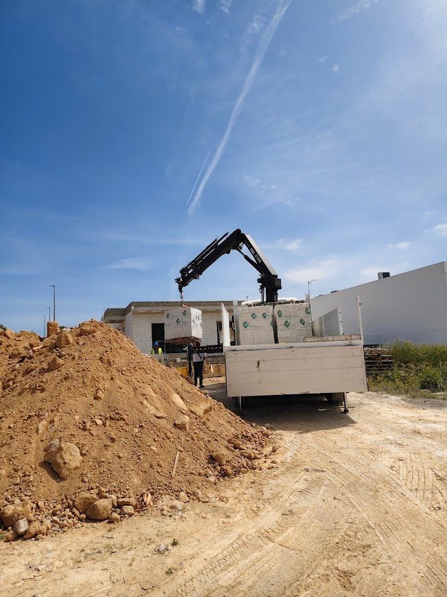 Obra en construcción con una excavadora cargando tierra junto a un camión volquete bajo un cielo azul despejado.