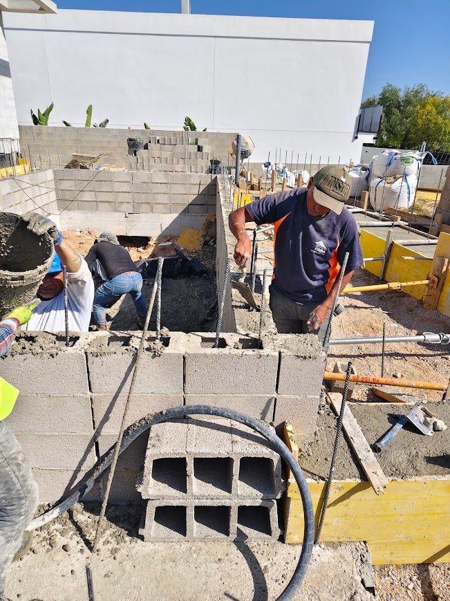 Obreros de la construcción colocando muros de bloques de hormigón en una obra, con herramientas y materiales en primer plano.