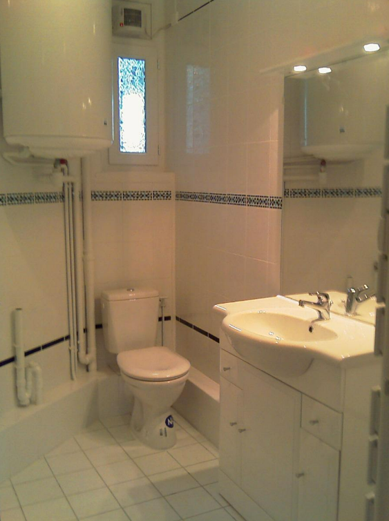 Une salle de bains blanche avec toilettes, lavabo et chauffe-eau. Des touches de bleu et une petite fenêtre.