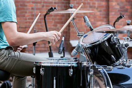 Un baterista toca una batería negra con baquetas de madera al aire libre, apoyado contra una pared de ladrillos.