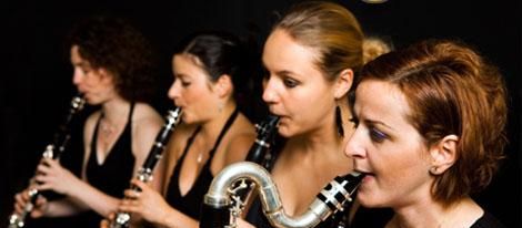 Cuatro músicos tocando clarinetes en fila, concentrados en su interpretación contra un fondo oscuro.