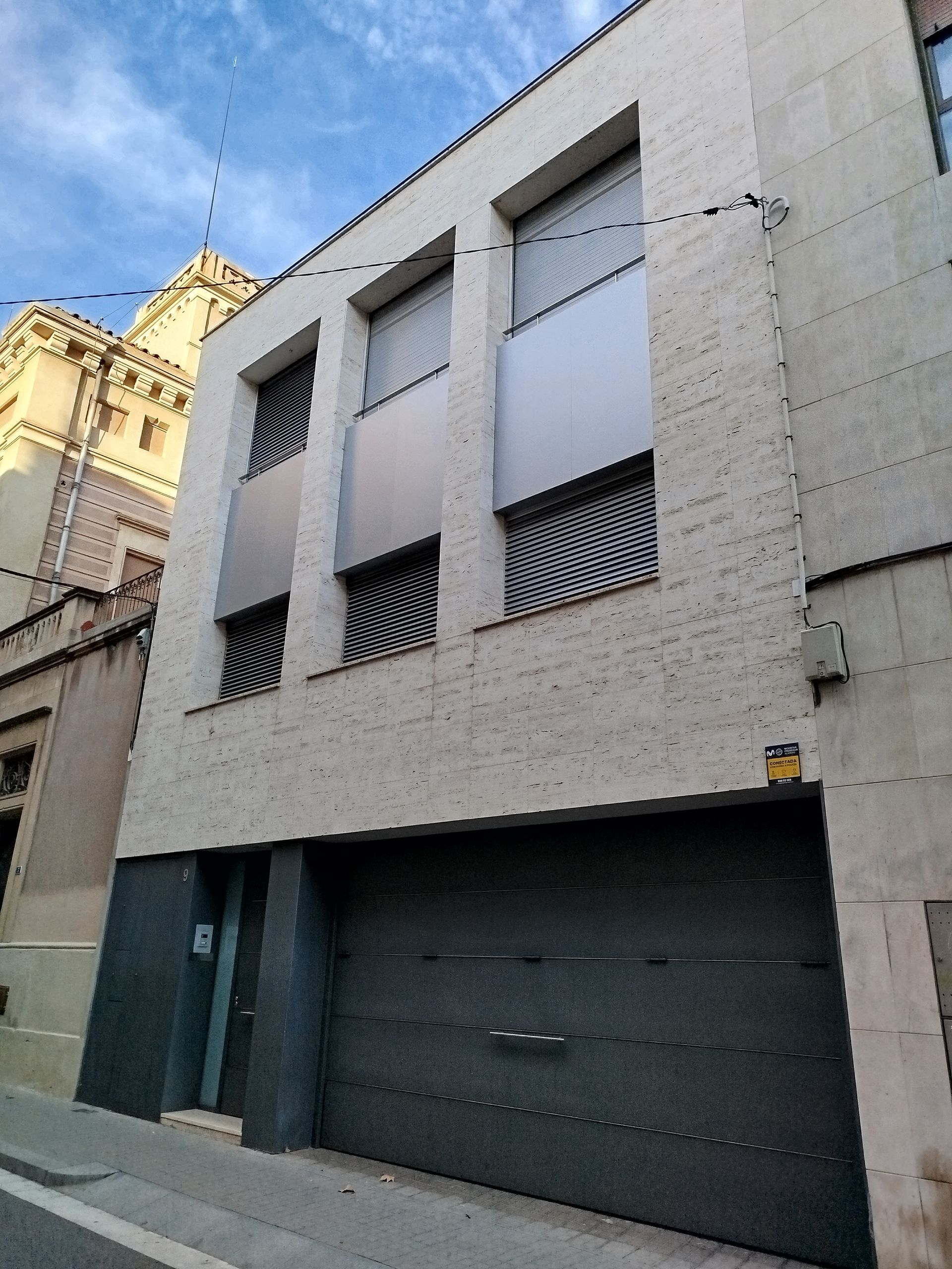 Edificio de tres plantas con puerta de garaje. Fachada gris con paneles metálicos y persianas.