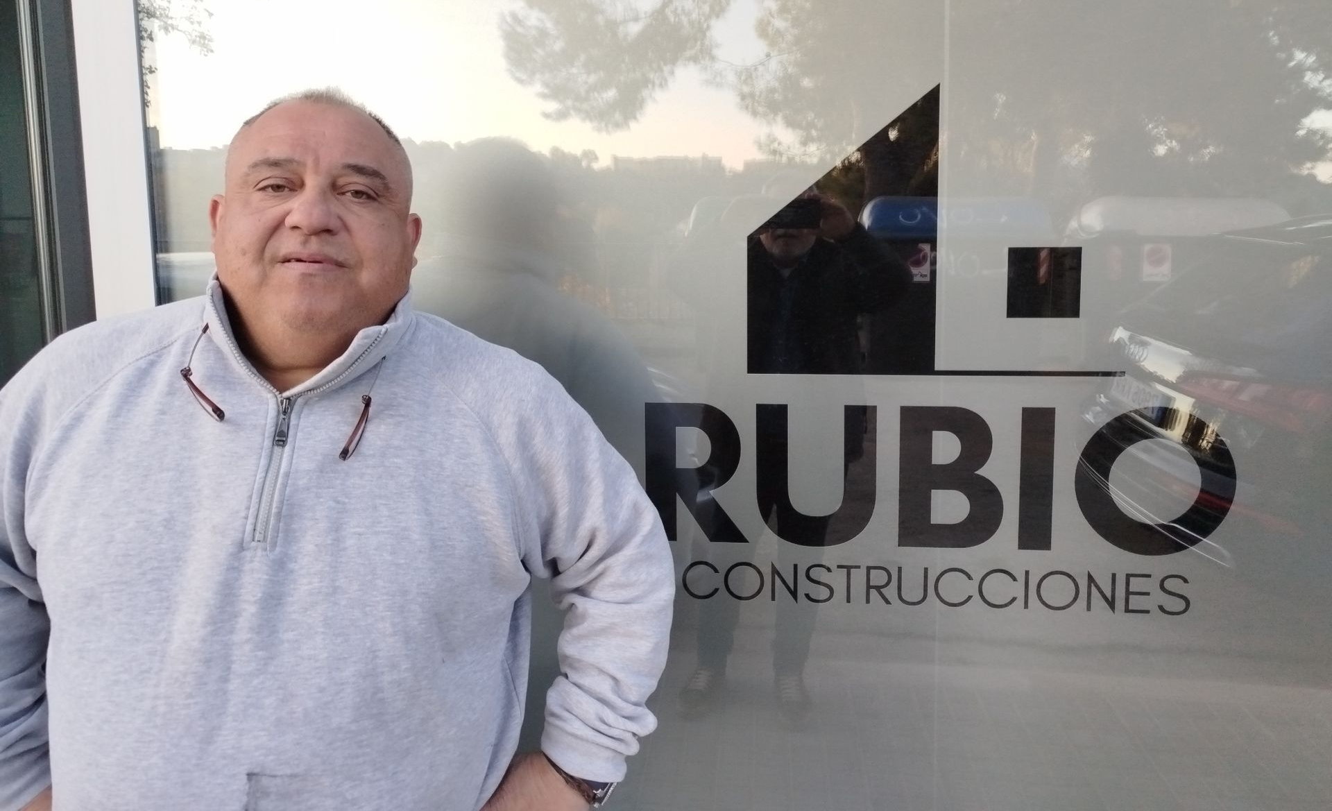 Hombre con camisa gris junto a una puerta de cristal con el logo de