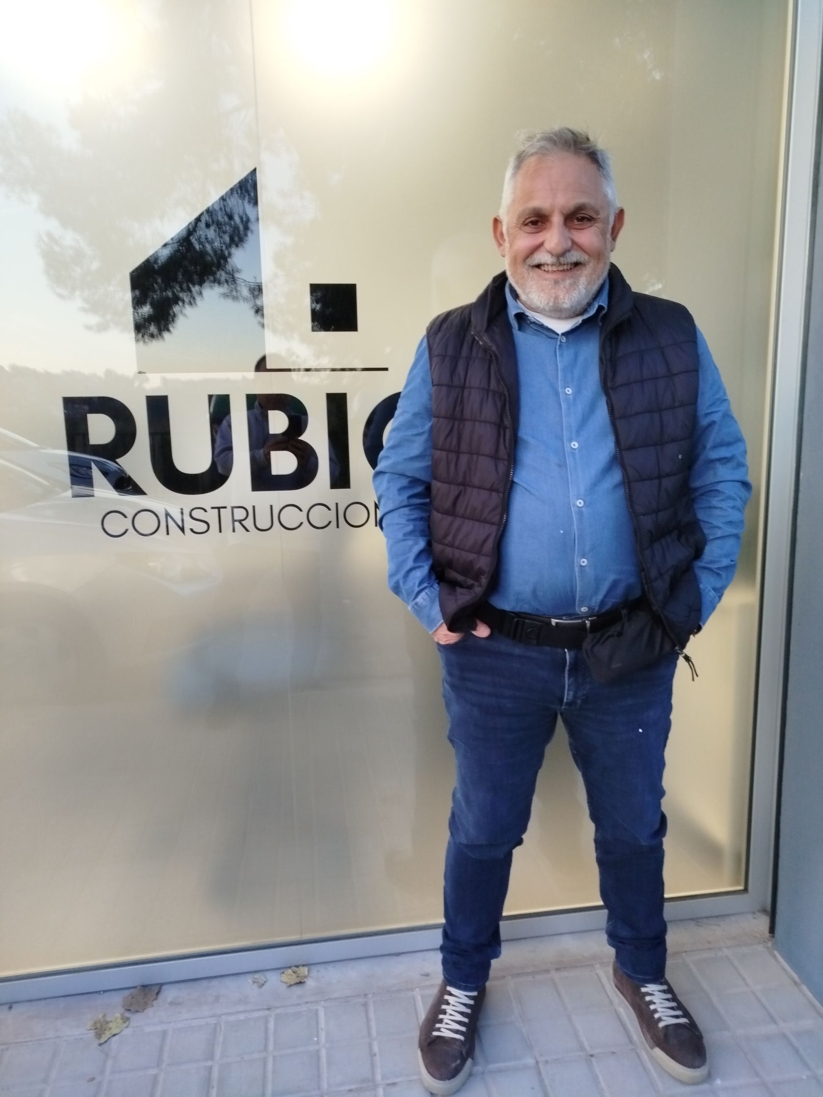 Hombre con camisa azul y chaleco frente al cartel de