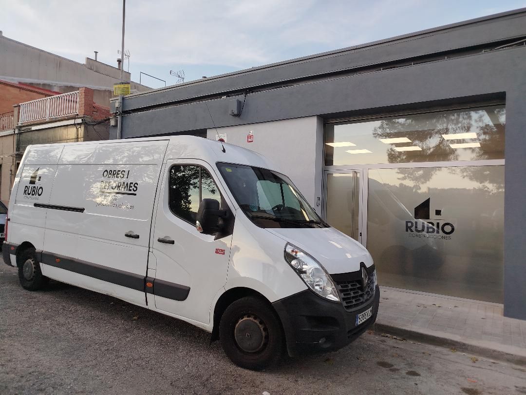 Una furgoneta blanca estacionada frente a un edificio con ventanas tintadas. La furgoneta tiene texto y logotipos