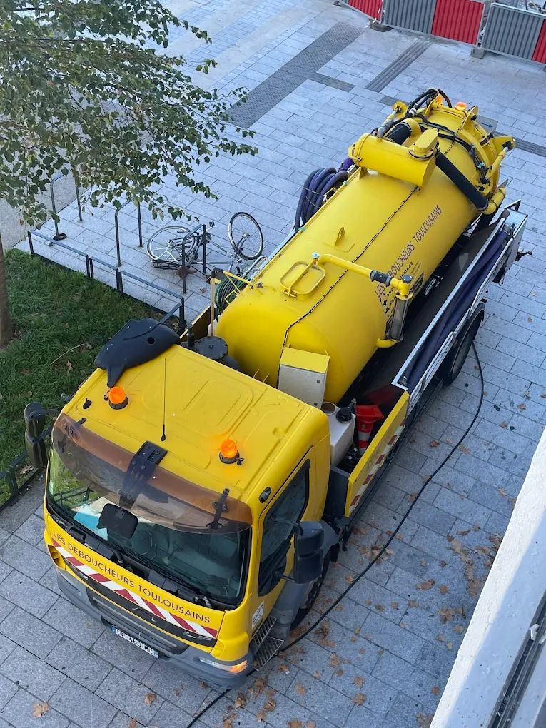 Un camion hydrocureur est stationné sur un trottoir pavé, à côté d'un bâtiment et d'arbres.