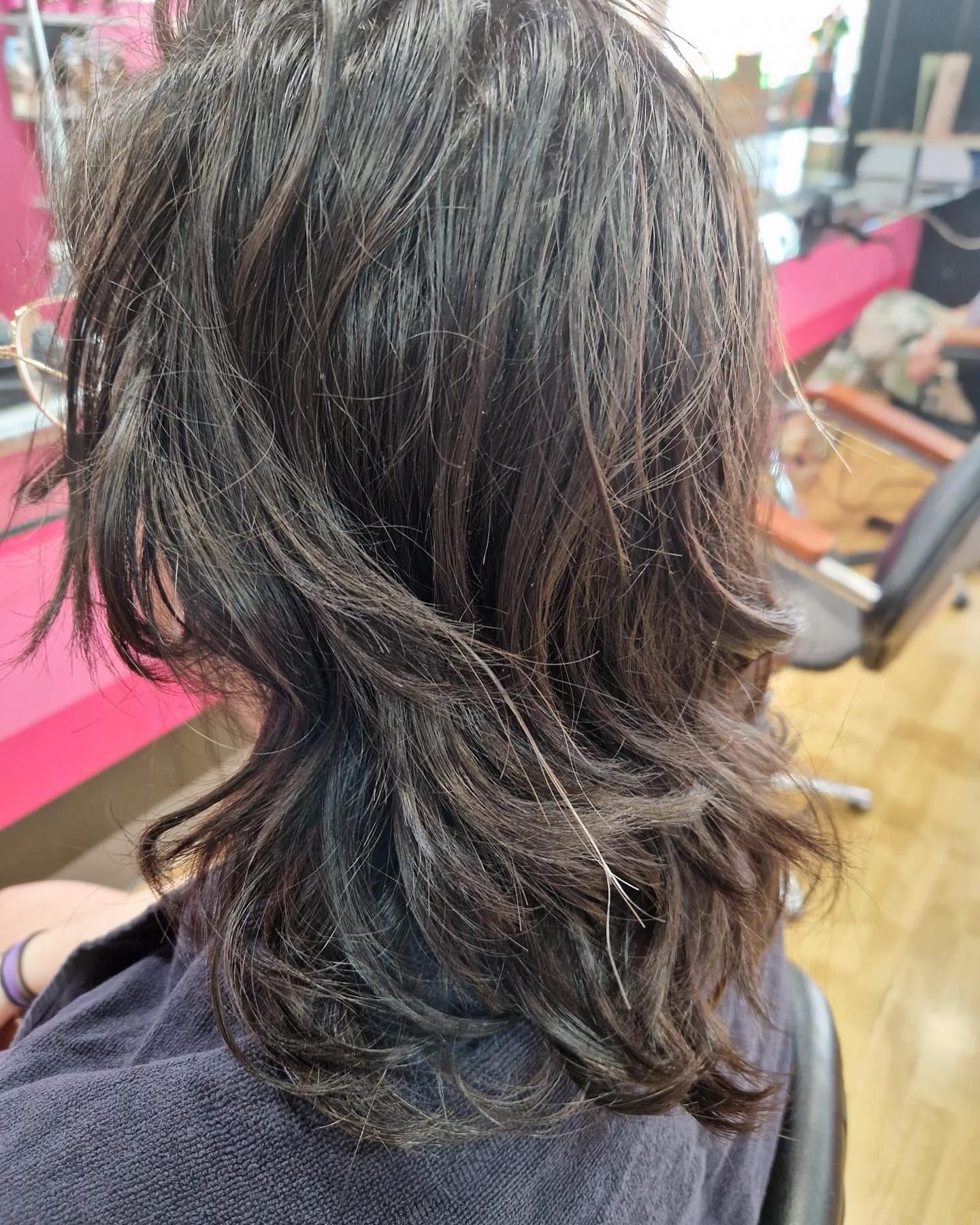 Vista posterior de una persona con cabello oscuro y en capas. Está sentada en una peluquería, posiblemente cortándose el pelo.