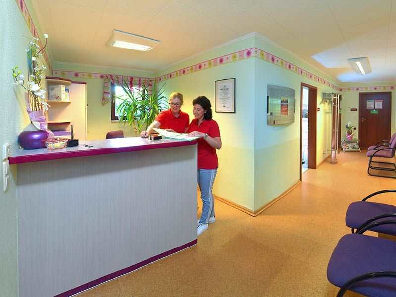 Zwei Personen an einem Empfangstresen in einer Arztpraxis; ein Wartezimmer ist sichtbar.