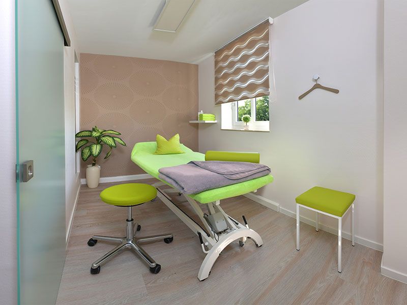 Massagetherapieraum mit grünen und grauen Möbeln, hellbraunen Wänden und einem Fenster.
