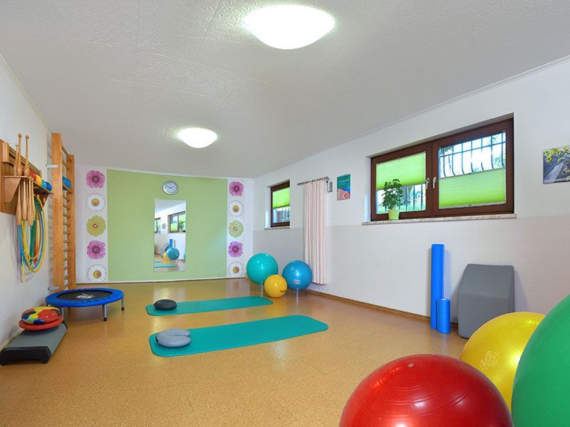 Fitnessraum mit Matten, Gymnastikbällen und Geräten; grüne und gelbe Wände; sonniger Raum.