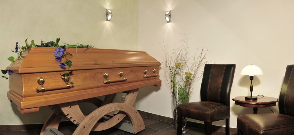 Sarg mit Blumen und Dekoration, rechts zwei leere Stühle.