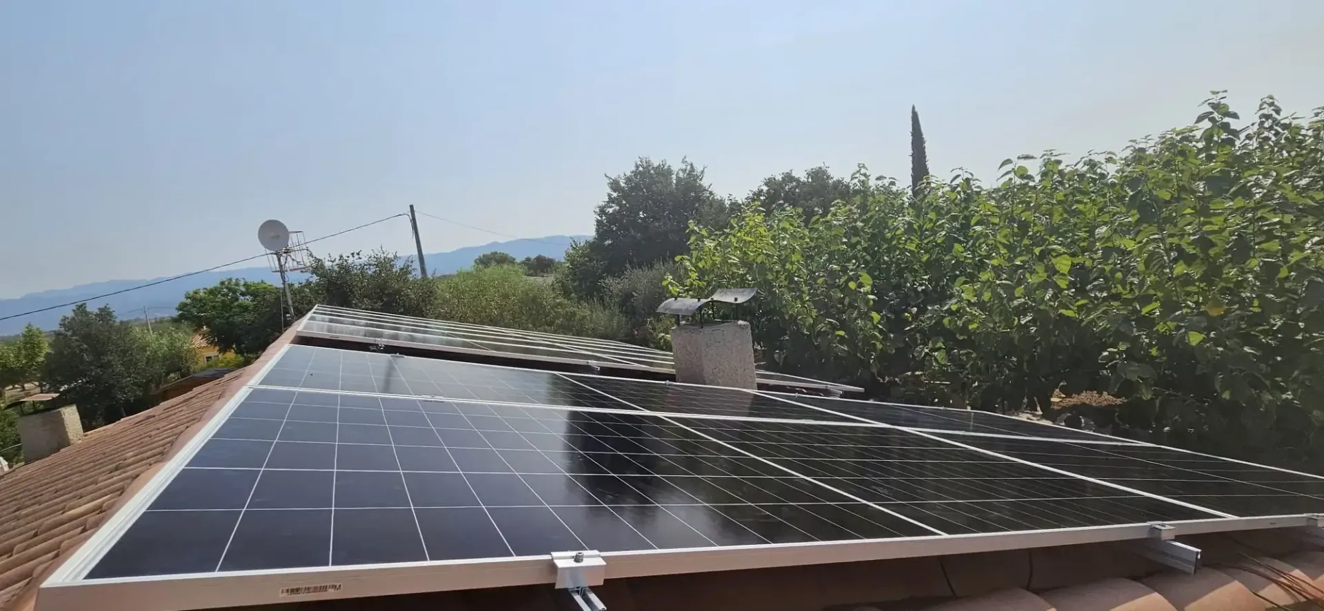 Paneles solares negros instalados en el tejado de tejas de una vivienda, rodeados de árboles bajo un cielo azul despejado.
