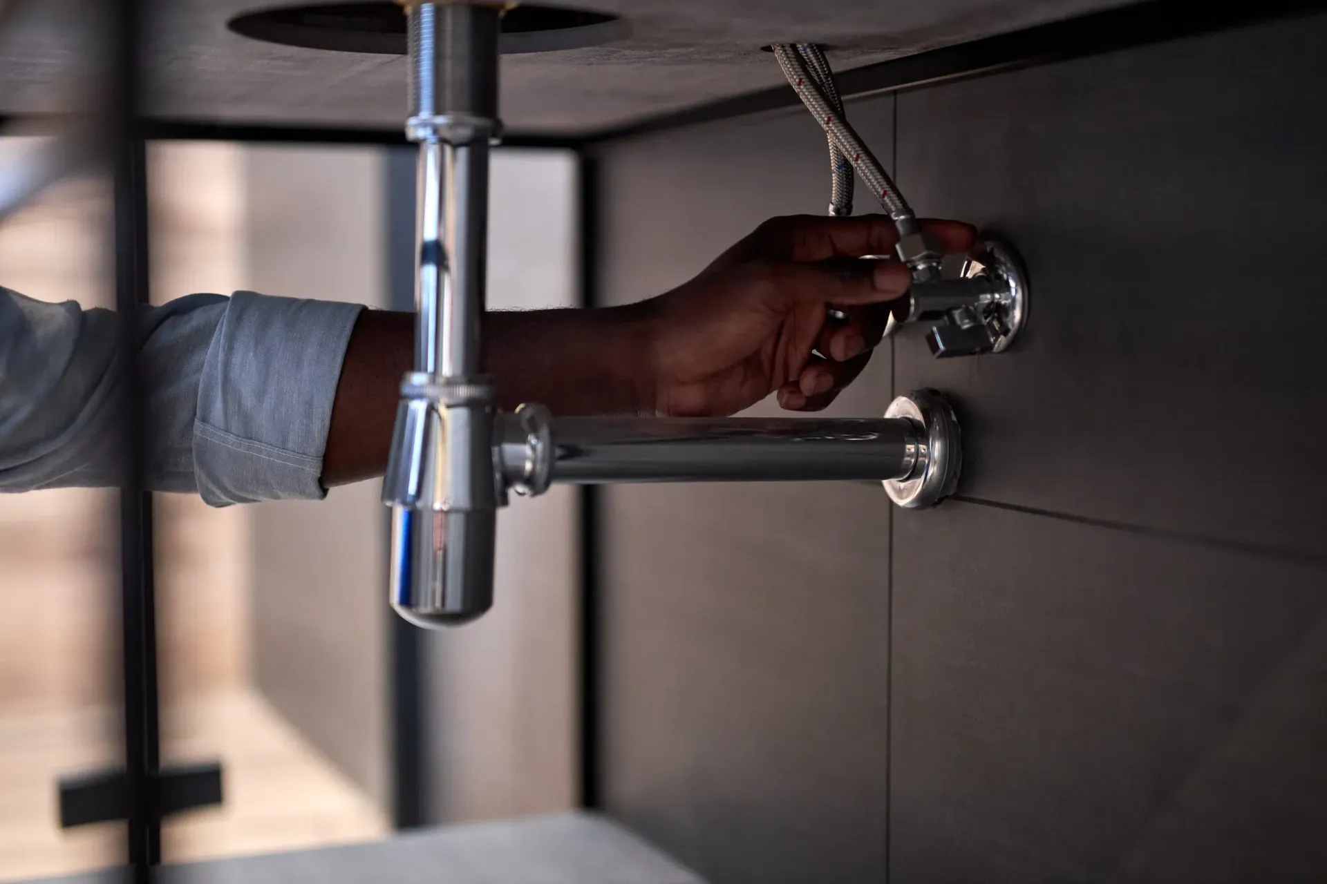 Primer plano de la mano de una persona ajustando un accesorio de plomería cromado debajo de un lavabo oscuro.