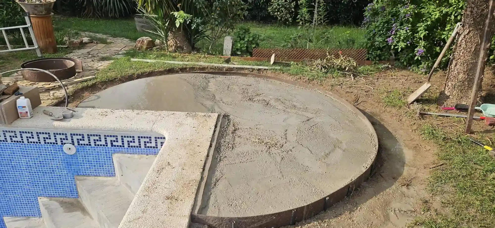 Un borde de patio circular de hormigón recién vertido junto a una piscina alicatada en un jardín trasero con césped.