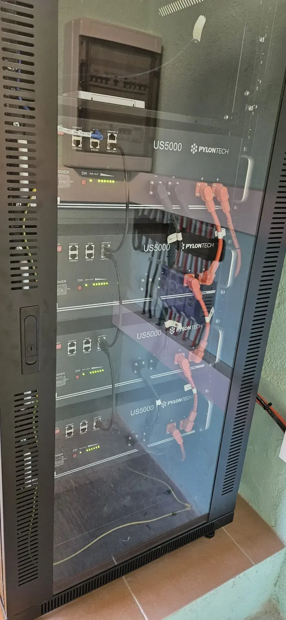 Un armario alto y oscuro para servidores, con puertas de cristal, que contiene equipos de red electrónicos y cables naranjas.