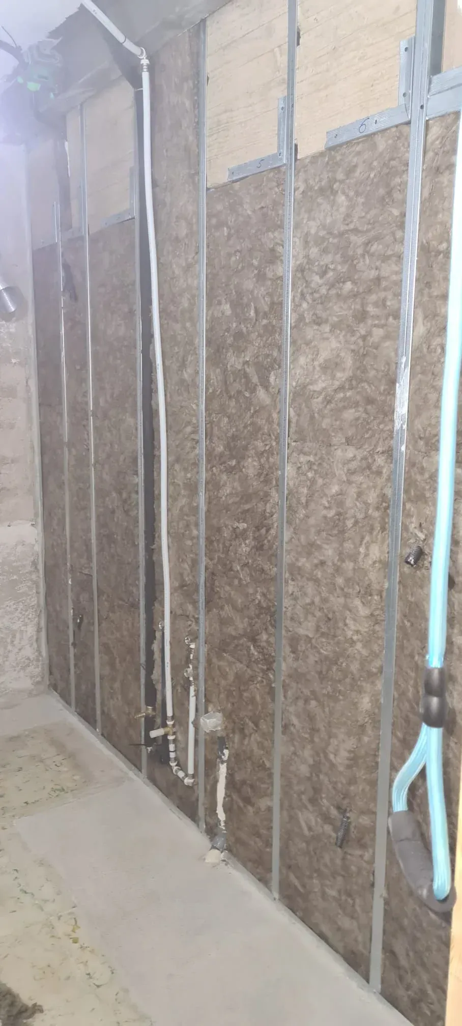 Vista de un muro en construcción con montantes metálicos, paneles aislantes y tuberías de fontanería a la vista.