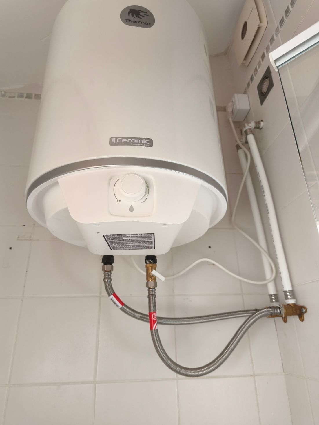 Calentador de agua blanco montado en la pared con tuberías a la vista en un rincón de baño alicatado.