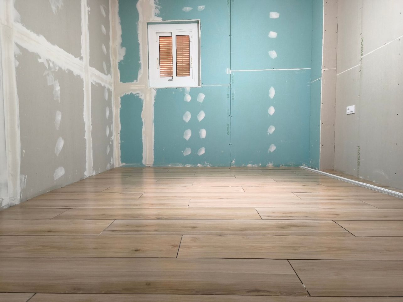 Habitación vacía en obras con paredes de pladur sin terminar, pared color turquesa y suelo de madera.