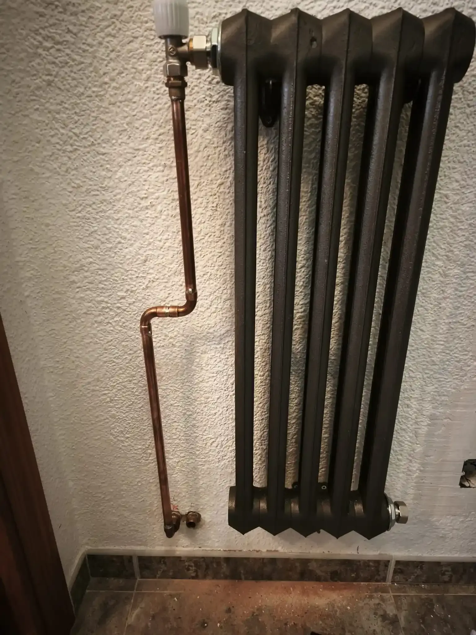 Un radiador mural de hierro fundido negro con tuberías de cobre conectado a una válvula blanca sobre una pared texturizada.