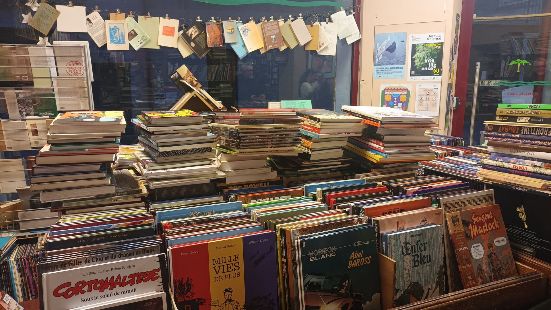Des piles de livres et de revues sont exposées dans une boutique, avec des cartes postales suspendues à une ficelle en arrière-plan.