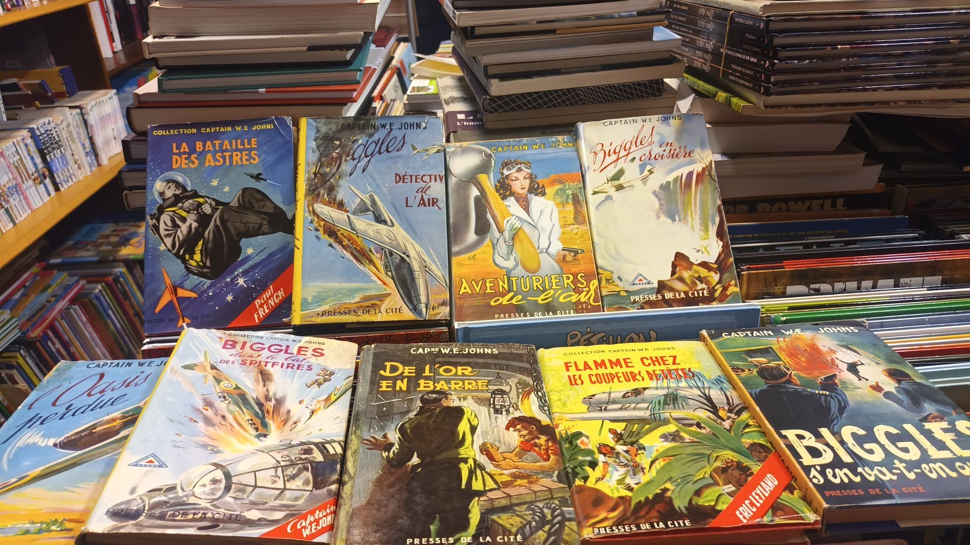 Vue en plongée de diverses couvertures de livres d'aventure anciens et de romans de Biggles exposées sur une étagère de librairie.