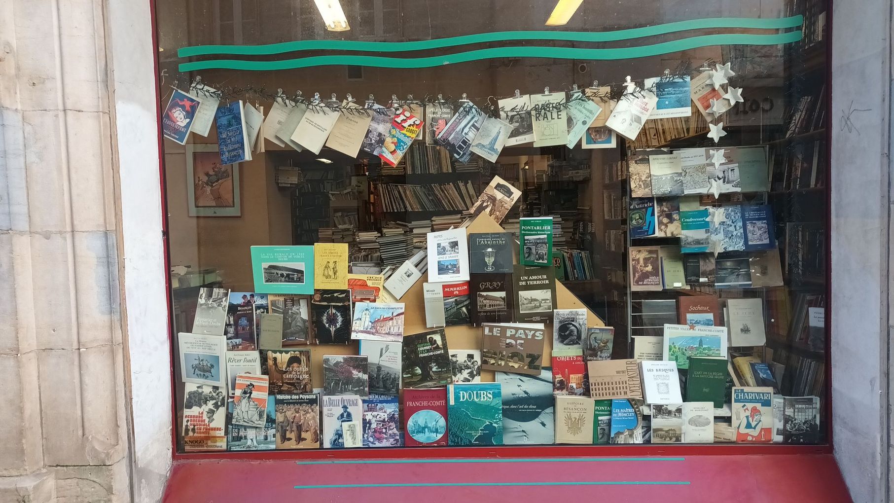 Une vitrine remplie d'une grande variété de livres et de cartes postales exposés, avec une bordure ondulée turquoise en haut.