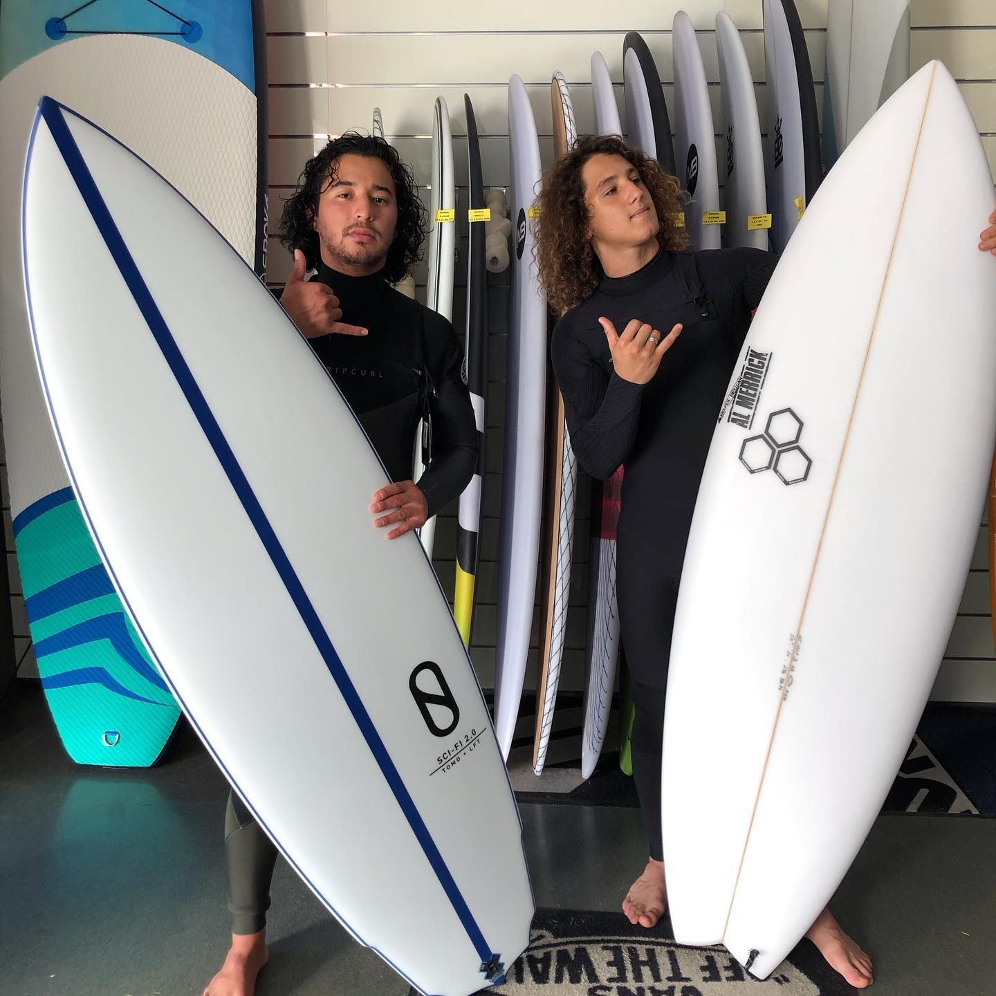 Deux personnes en combinaisons de surf, tenant des planches de surf, font le signe shaka à l'intérieur d'un magasin de surf.