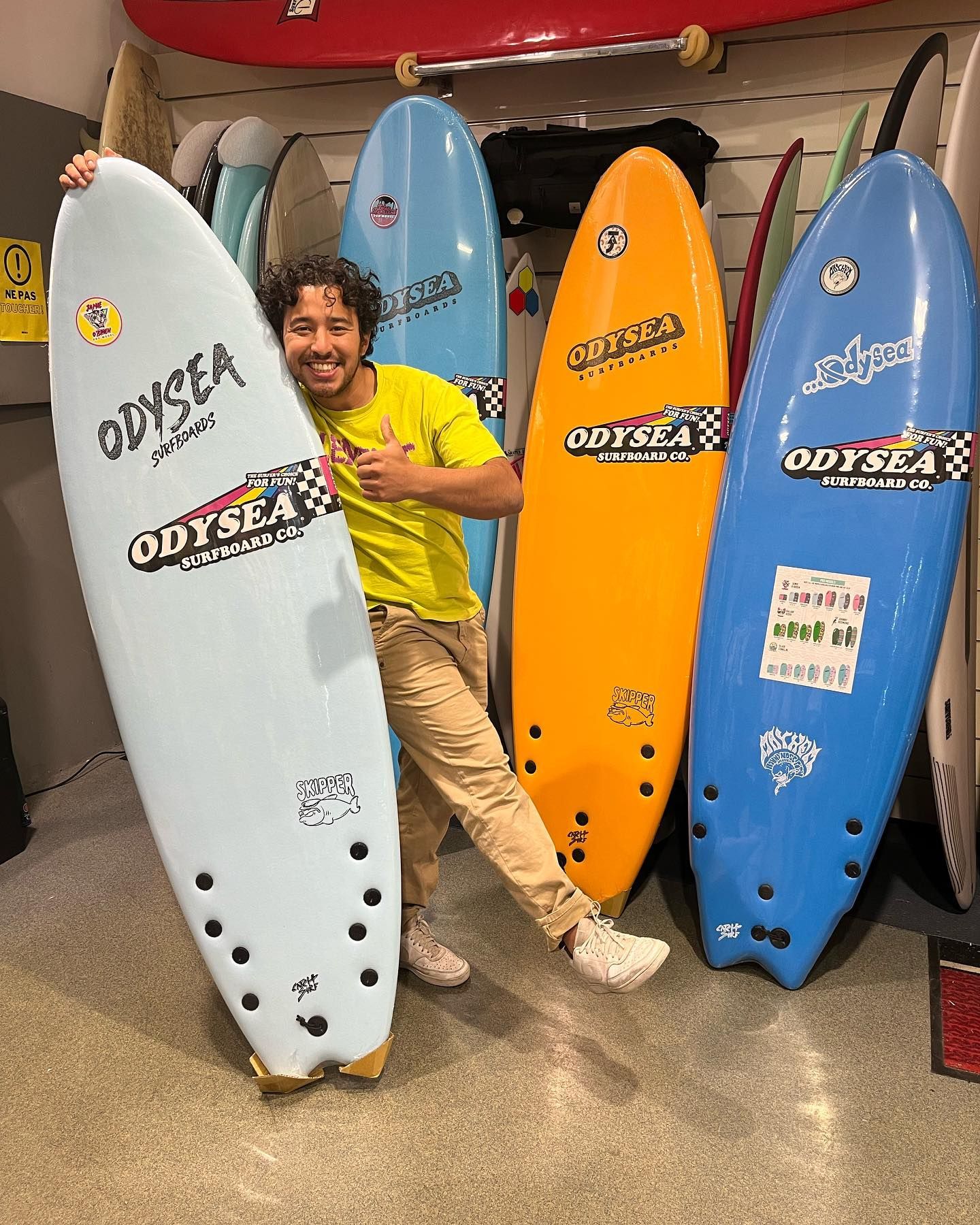 Un homme sourit, pouce levé, et pose avec des planches de surf colorées dans un magasin.