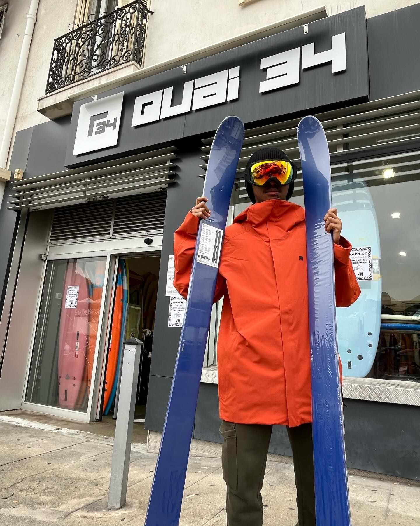 Une personne en veste orange tenant des skis devant un magasin portant l'inscription « Ques 34 ».