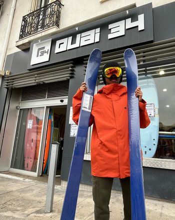 Une personne tenant des skis devant le magasin « Quai 34 ». Elle porte une veste orange, des lunettes de ski et sourit.